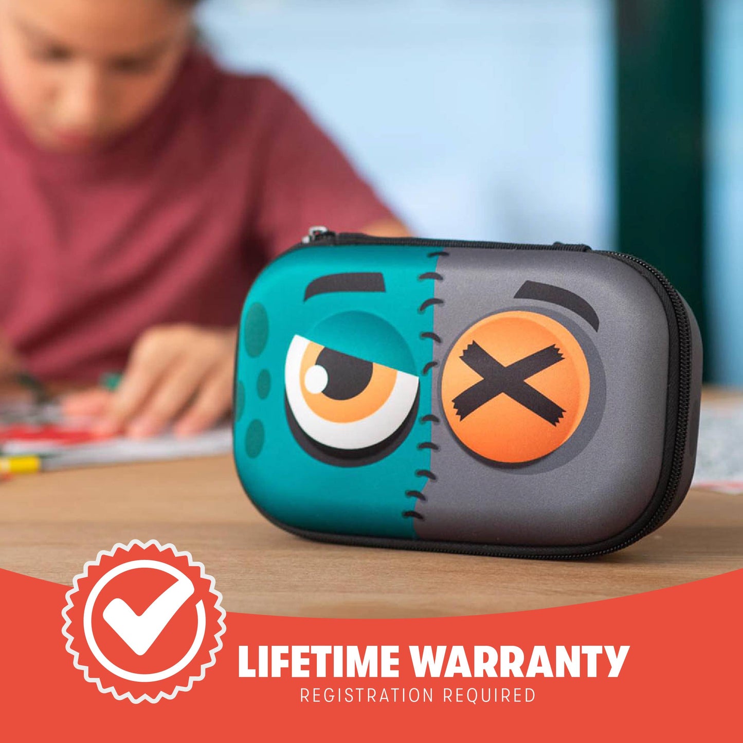 Children's pencil case with cartoon face design on a table, with a child writing in the background. Lifetime warranty badge visible.